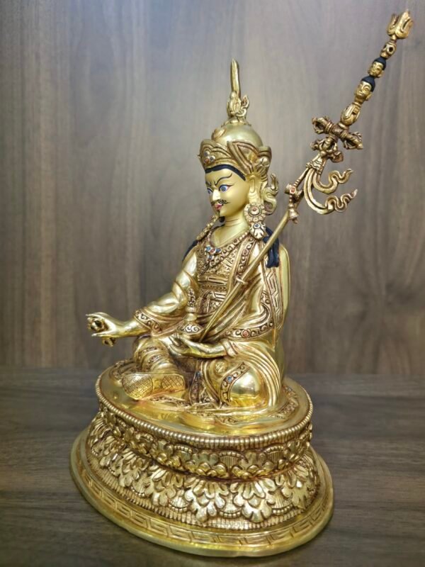 Side angle of Guru Rinpoche statue showing the khatvanga trident staff resting against the left shoulder.