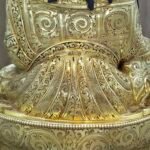 Close up back view of gold plated Guru Rinpoche statue showing intricate floral engravings on the robes and flowing blue hair.