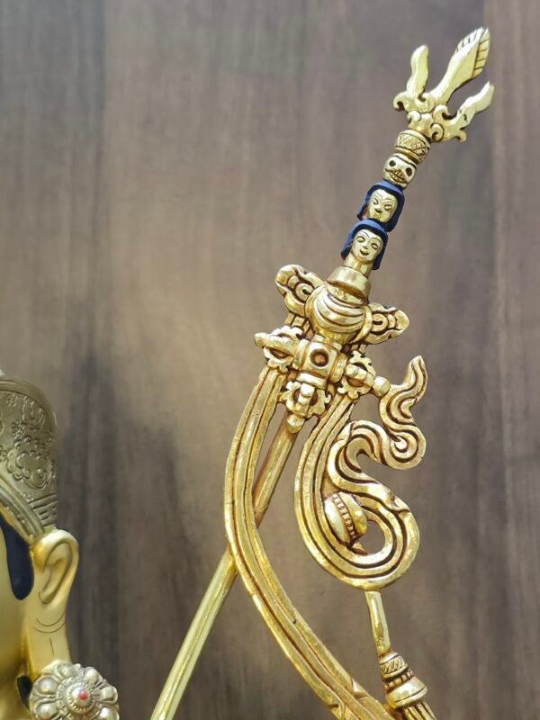 Detail of Khatvanga trident staff on Padmasambhava statue, featuring heads and hanging sash ornaments in gold.