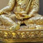 Detail of Guru Rinpoche statue hand holding the Tse Bum Long Life Vase and Vajra, showing intricate gold plated patterns on the robes.