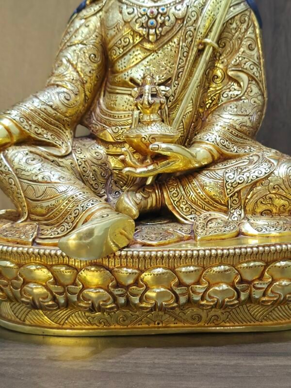 Detail of Guru Rinpoche statue hand holding the Tse Bum Long Life Vase and Vajra, showing intricate gold plated patterns on the robes.