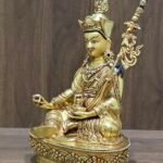 Angled view of Guru Rinpoche statue showing gold plated finish, Khatvanga staff, and gem-set jewellery.