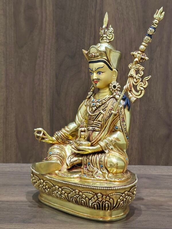 Angled view of Guru Rinpoche statue showing gold plated finish, Khatvanga staff, and gem-set jewellery.