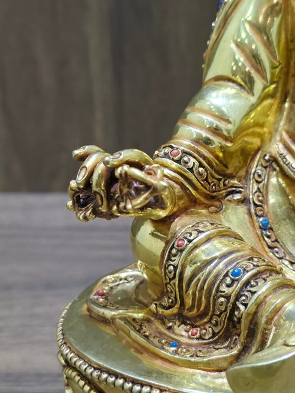 Close up detail of Guru Rinpoche right hand holding gold Vajra, showing hand-chased floral patterns on robes and gem settings.