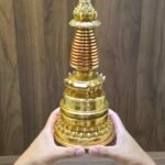 Gold plated copper Buddhist stupa held in two hands for scale, showing red spire rings and lotus base.
