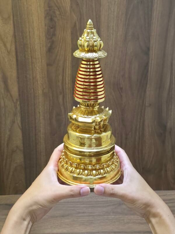 Gold plated copper Buddhist stupa held in two hands for scale, showing red spire rings and lotus base.
