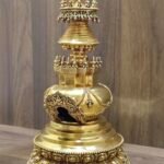 Angled left view of gold plated copper stupa showing torana arch window, rounded body, and ringed spire under jeweled canopy.