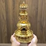 Stupa held in hands for scale, showing gold plated copper finish, carved lotus base, and ornate arch window on front.