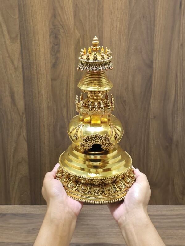 Stupa held in hands for scale, showing gold plated copper finish, carved lotus base, and ornate arch window on front.