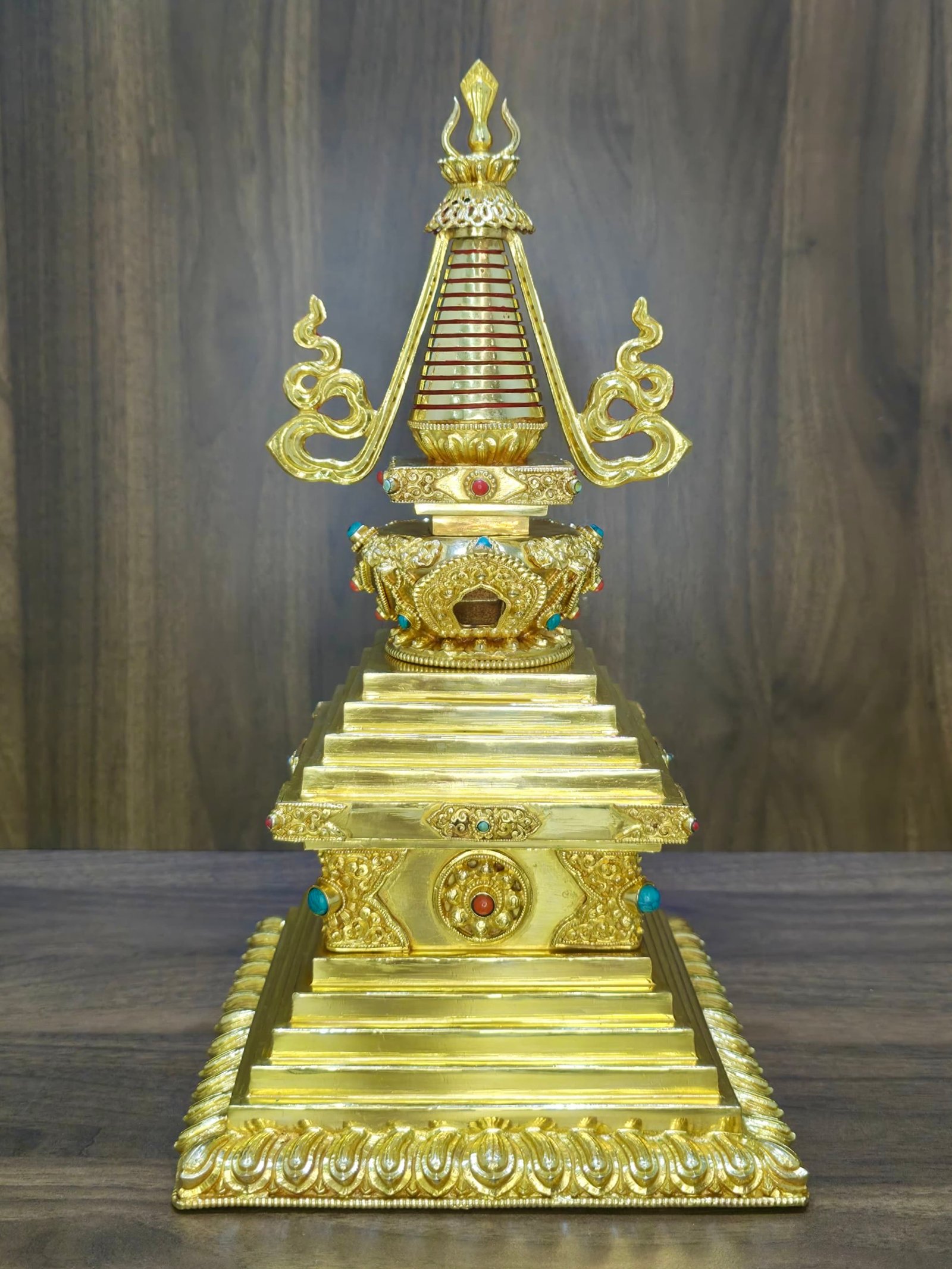 Gold plated copper Tibetan style stupa front view on wooden backdrop, tiered square base with lotus border and ringed spire.