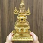 Hands holding the stupa to show size and scale, gold plated finish with stepped square base and tall spire.
