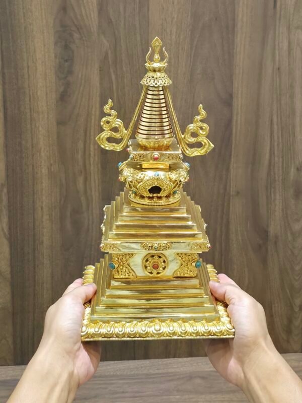 Hands holding the stupa to show size and scale, gold plated finish with stepped square base and tall spire.