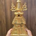 Handheld view of gold plated copper Tibetan style stupa with tiered square base and curved side ornaments beside the spire.