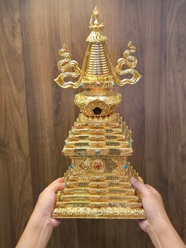 Handheld view of gold plated copper Tibetan style stupa with tiered square base and curved side ornaments beside the spire.
