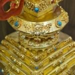 Close up of the stupa’s stepped base showing engraved panels, raised floral trim, and small blue and red inlay accents.