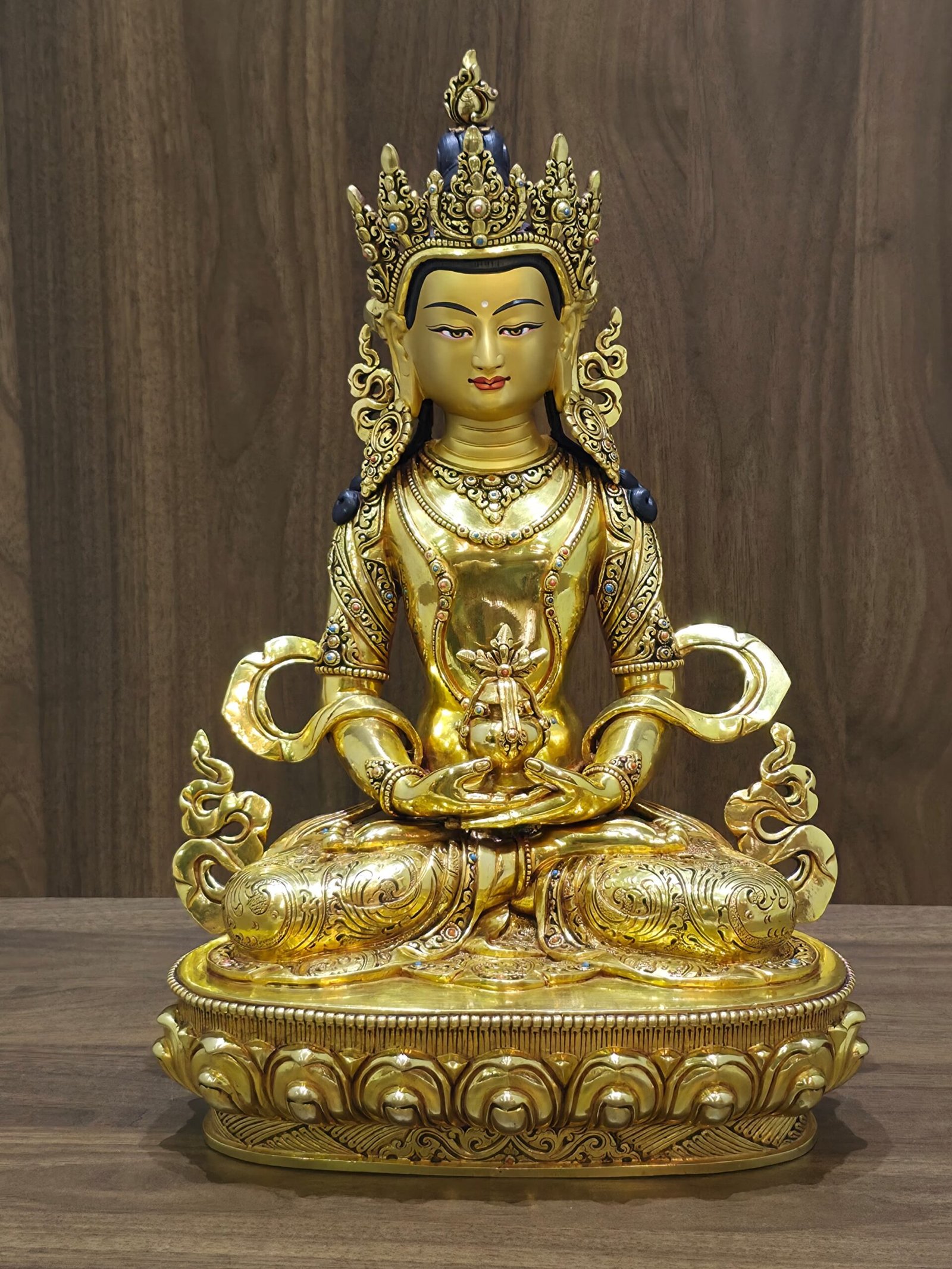 Front view of gold plated Amitayus (Aparimita) Buddha seated on a lotus pedestal, holding a long-life vase beneath an ornate crown.