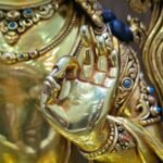 Close-up of Manjushri’s raised left hand, with thumb and forefinger touching, beside an ornate bracelet and necklace.