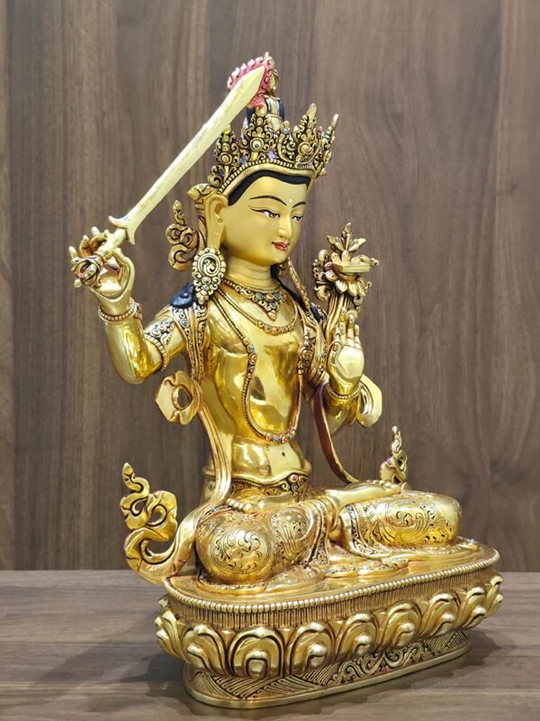 Manjushri statue angled to the right, showing the sword handle, raised hand, flowing scarf and engraved lotus pedestal.
