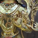 Close-up of Manjushri’s raised left hand, with thumb and forefinger touching, beside layered necklaces and a decorated bracelet.