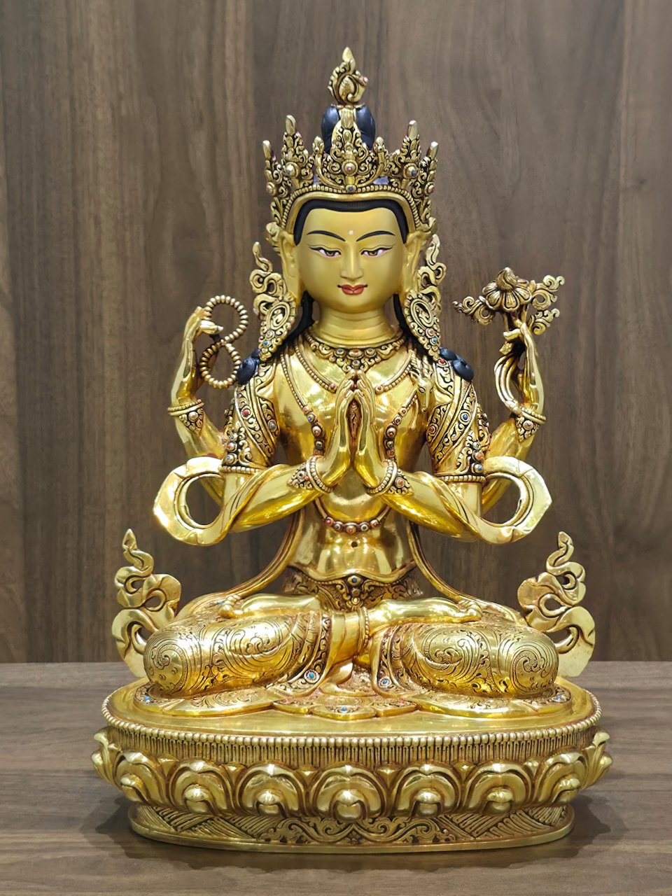 Full frontal view of four-armed Chenrezig (Avalokiteshvara) statue seated on a lotus pedestal against a wood background.