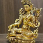 Green Tara statue facing left, showing the open right palm, lotus foot support and decorated pedestal against a wood background.