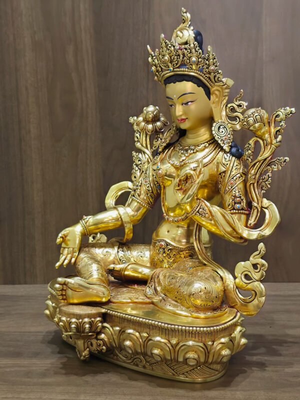 Green Tara statue facing left, showing the open right palm, lotus foot support and decorated pedestal against a wood background.