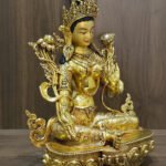 Green Tara statue facing right, showing the shoulder flowers, flowing scarf ornaments and engraved garments against a wood background.