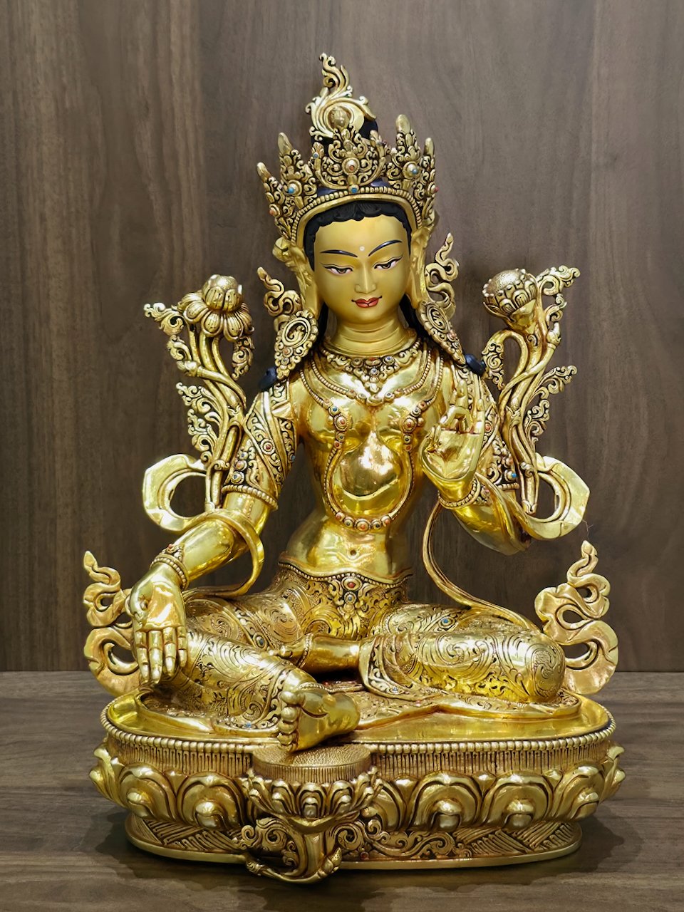 Front view of a 24K gold plated copper Green Tara statue seated on a lotus pedestal against a wood background.