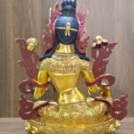 Rear view of the gold plated Green Tara statue showing dark painted hair, engraved garments and lotus base against a wood background.