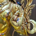 Seven-Eyed White Tara statue in 24K gold plated copper, close-up of the painted eye on the raised left palm beside a lotus stem.
